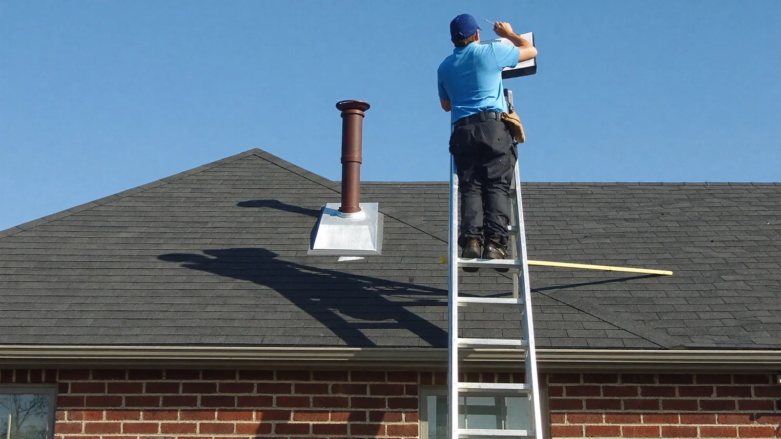 Roof inspector on a ladder at the eave of a shingle roof photographing flashing with a tablet, a one-story Tennessee home below