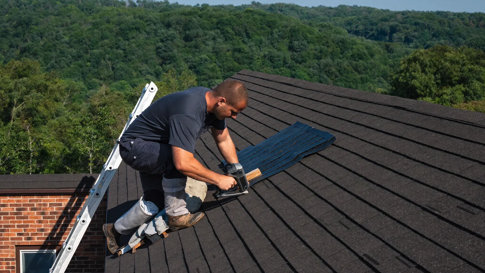 Roofer kneeling on a steep asphalt shingle roof nailing down a course of architectural shingles on a two-story brick home in Clarksville, Tennessee, under a clearing sky