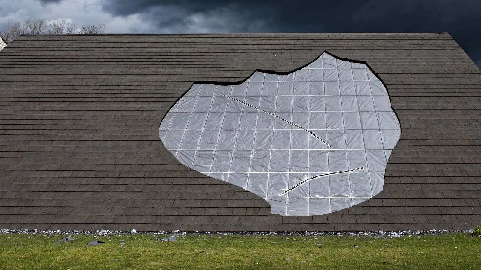 Residential roof with a patch of shingles torn away by wind, exposing underlayment and nail heads, under a dark Middle Tennessee storm sky
